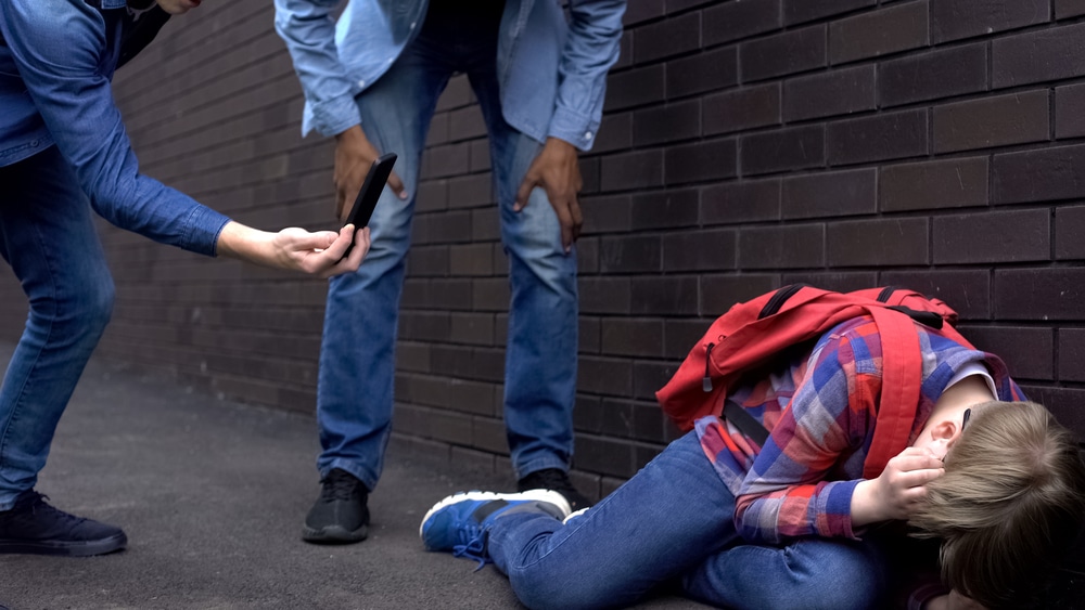 Une personne accroupie contre un mur se couvrant la tête tandis qu'une autre personne ramasse un téléphone portable tombé à proximité d'un agresseur potentiel, tous deux impliqués dans la lutte contre la haine en ligne.